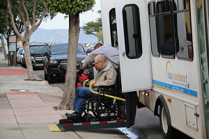 old man in a wheelchair is assisted to get off a van using a lift for senior transportation service